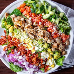 Cobb Salad is a classic American dish that typically includes chopped salad greens, tomato, crispy bacon, grilled chicken, hard-boiled eggs, avocado, chives, Roquefort cheese, and a red-wine vinaigrette.