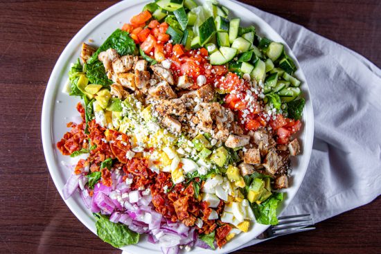 Cobb Salad is a classic American dish that typically includes chopped salad greens, tomato, crispy bacon, grilled chicken, hard-boiled eggs, avocado, chives, Roquefort cheese, and a red-wine vinaigrette.